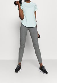 Person som bär en ljusblå T-shirt och grå leggings, lyfter svarta hantlar med böjda armar, står mot en enkel vit bakgrund.