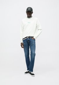 Λευκό φούτερ με τιρκουάζ λογότυπο, μαύρο γιακά, και μπλε τζιν. Μαύρο καπέλο και αθλητικά παπούτσια ολοκληρώνουν το καθημερινό ντύσιμο.