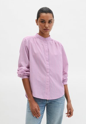 Femme aux cheveux tirés en arrière, portant une chemise à manches longues rayée verticalement rose et blanche et un jean bleu clair, posant devant un fond uni.