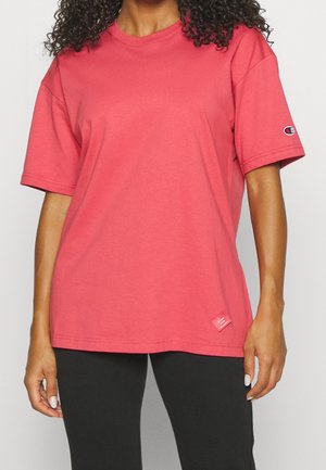 Kobieta w luźnej, koralowo-różowej koszulce z krótkim rękawem z małą naszywką z logo na rękawie i na dole, w połączeniu z czarnymi spodniami.