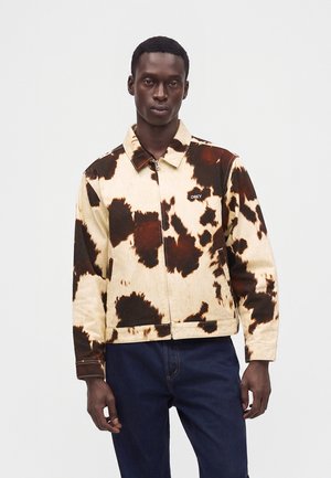 Mand iført en lynlåstrøje med et brun og cremefarvet ko-mønster og mørkeblå jeans, stående mod en ensfarvet baggrund.