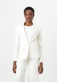 Weißer Leinenblazer mit Reverskragen, einreihiger Knopfschluss und zwei Vorderitaschen. Kombiniert mit einem passenden weißen Oberteil und einer Hose.