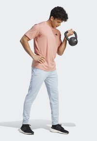 Rosa trænings-T-shirt med logo, lyseblå tapered bukser og sorte sneakers. Holder en sort kettlebell med mat finish.