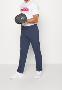 Marine joggingsbroeken van zacht materiaal, met een elastische tailleband. Gecombineerd met een wit T-shirt en witte sportschoenen, terwijl hij een gewicht vast heeft.