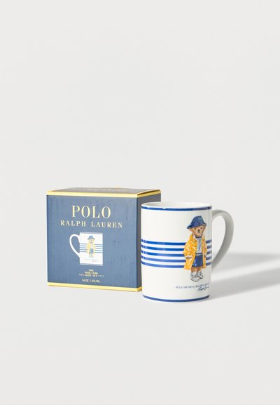 Fehér kerámia bögre kék csíkokkal, mellett egy medve, aki sárga esőkabátot és kék kalapot visel, és egy márkázott tengerészkék és arany színű doboz.