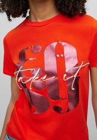 Czerwona bawełniana koszulka z wyrazistym metalicznym srebrno-białym napisem "60 take it" na przodzie. Krótkie rękawy i okrągły dekolt.