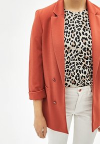 Červené dvouřadé sako s ohrnutými rukávy přes béžový top s černobílým leopardím potiskem, doplněné bílými kalhotami.