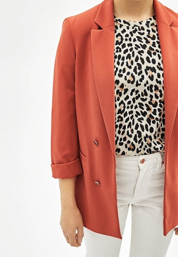 Červené dvouřadé sako s ohrnutými rukávy přes béžový top s černobílým leopardím potiskem, doplněné bílými kalhotami.