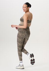 Glunderende zwangere vrouw in een actieve outfit met luipaardprint, witte sokken en grijze New Balance-sneakers, poserend met één voet iets omhoog.
