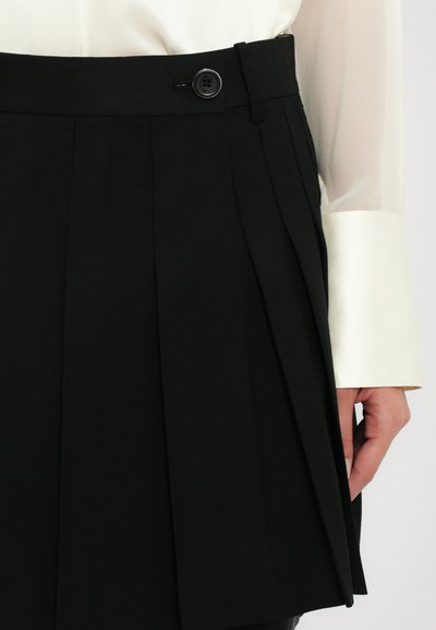 Gonna nera plissettata con vita alta, texture liscia e chiusura con bottone nella parte anteriore, abbinata a una blusa bianca in satin con maniche.