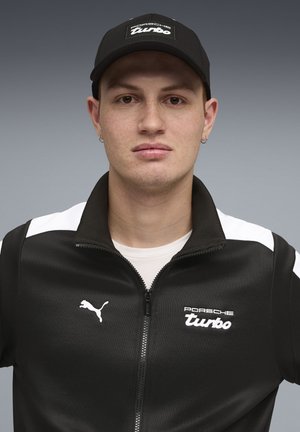 Zwarte jas met ritssluiting en witte accenten; met Puma-logo en tekst "Porsche Turbo". Draagt bijpassende zwarte pet. Gladde textuur.