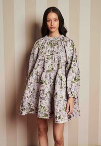 Žena s dlhými tmavými vlasmi, oblečená v voľných šatách s kvetinovým vzorom a nadúvanými dlhými rukávmi, stojí proti béžovej pruhovanej stene.