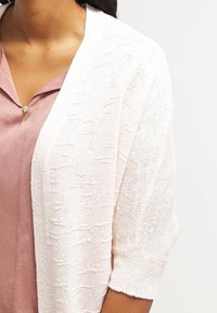 Světle růžový pletený kardigan s krátkými rukávy, který má texturovaný vzor a otevřený přední design. Měkká a prodyšná tkanina.