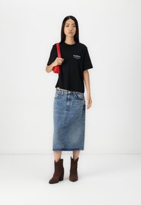 Camiseta negra de manga corta con logo blanco, combinada con una falda midi de mezclilla azul y botas de vaquero marrones de tacón. Un bolso cruzado rojo acentúa el look.
