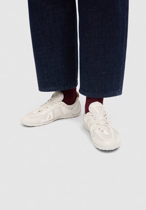 Personne portant un jean large bleu foncé, des chaussettes bordeaux et des baskets à lacets blanc cassé, debout sur un sol gris clair.
