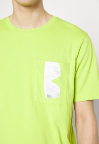 Personne portant un t-shirt vert citron avec une poche poitrine ornée d'un design abstrait blanc et argenté brillant ressemblant à des lunettes de soleil.