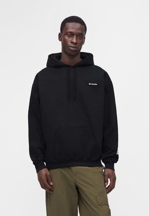 Homme portant un sweat à capuche noir Columbia avec poche avant et pantalon cargo vert olive sur un fond clair uni.