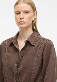 Smeđa traper jakna s ovratnikom, opremljena crnim metalnim gumbima i vertikalnim šavnim detaljima. Glatka tekstura, ležeran dizajn.
