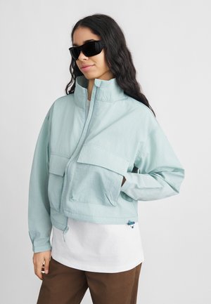 Femeie purtând ochelari de soare negri, o jachetă albastru deschis cu fermoar și buzunare mari în față, cămașă albă și pantaloni maro, stând cu o mână în buzunar.