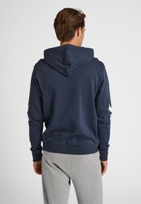 Marinblå bomulls-hoodie med dragsko i huvan, ribbstickade mudd och en kontrasterande vit designdetalj längs ärmarna. Mjuk textur, avslappnad passform.