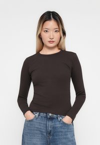 Kvinne med langt hår som har på seg en tettsittende mørkebrun langermet topp og blå jeans, med hendene i lommene, vendt mot kameraet foran en ensfarget bakgrunn.