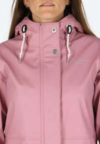 Giacca impermeabile rosa con colletto alto, lacci bianchi e bottoni a pressione argentati. Presenta un logo sul petto e una texture liscia.