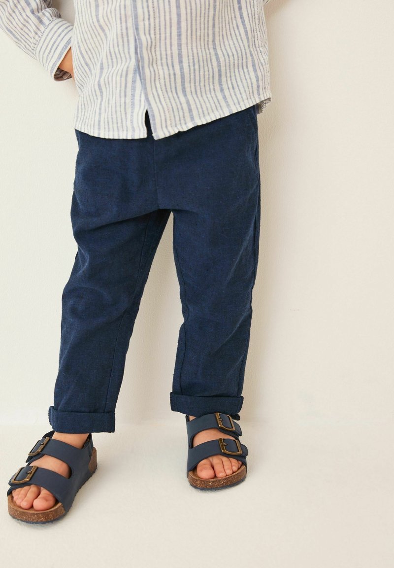 Enfant portant un pantalon bleu marine retroussé et des sandales bleu marine à boucles doubles, debout devant un fond clair uni.