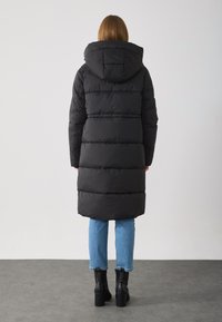 Pardessus nero imbottito con cappuccio, caratterizzato da un design trapuntato e una lunghezza fino al ginocchio. Indossato sopra un paio di jeans blu chiaro a vita alta e stivali neri con tacco.