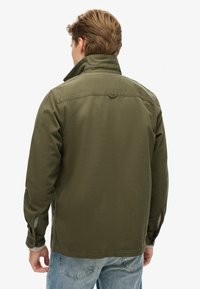 Chaqueta verde oliva con un cuello estructurado, que presenta un yugo trasero y detalles en los puños. Está hecha de una tela resistente, mostrando una textura lisa.