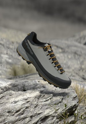 Chaussure de randonnée La Sportiva grise et noire avec des lacets jaunes flottant au-dessus d'un terrain rocheux avec de petites plantes.
