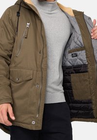 Veste isolante vert olive avec fermeture éclair sur le devant, poche poitrine et capuche doublée de fourrure ; dotée d'une doublure matelassée grise et d'une poche intérieure noire.