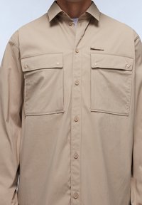 Camicia beige con bottoni, realizzata in tessuto leggero, con due tasche sul petto, colletto classico e texture liscia con un leggero brillantezza.