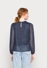 Blusa azul-marinho com tecido transparente riscado verticalmente, mangas bufantes e fecho de botão nas costas; combinada com calças de denim claro.