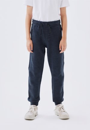 Dziecko ubrane w ciemnoniebieskie spodnie dresowe ze sznurkiem w talii i białe trampki, stojące na jednolitym tle.