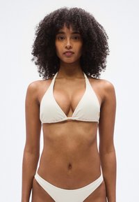 Τριγωνικό σετ μπικίνι σε ανοιχτό κρεμ υφή με ριμπ, με λεπτές τιράντες και ελάχιστη κάλυψη. Περιλαμβάνει μικρή διακόσμηση λογότυπου στην κορυφή.