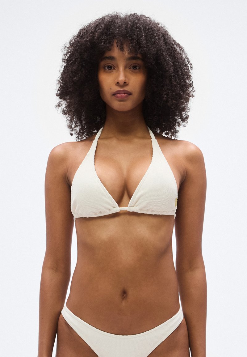 Τριγωνικό σετ μπικίνι σε ανοιχτό κρεμ υφή με ριμπ, με λεπτές τιράντες και ελάχιστη κάλυψη. Περιλαμβάνει μικρή διακόσμηση λογότυπου στην κορυφή.