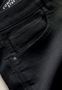 Čierna denimová látka s hladkou textúrou má zadnú vrecko, obrysové stehy a kovový detail gombíka. Na etikete je napísané "Comfort Stretch."