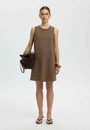 Femme portant une robe marron sans manches tenant un sac à main tressé, portant des boucles d'oreilles anneaux, des bracelets et des sandales noires avec anneaux pour orteils.