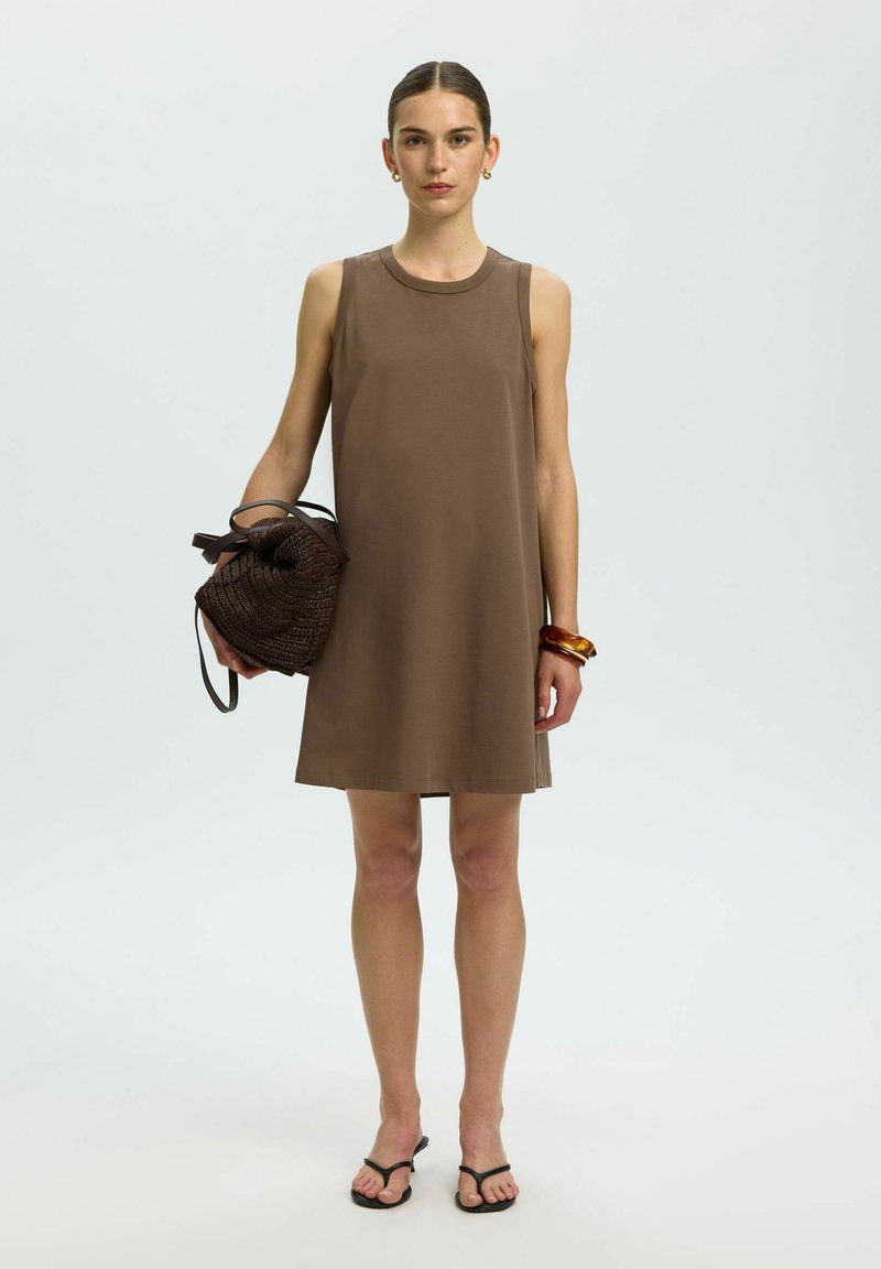 Femme portant une robe marron sans manches tenant un sac à main tressé, portant des boucles d'oreilles anneaux, des bracelets et des sandales noires avec anneaux pour orteils.