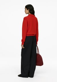 Roter Strickpullover mit Kragen, kombiniert mit weiten schwarzen Hosen. Trägt eine runde, bordeauxfarbene Wildledertasche. Einfacher Hintergrund.