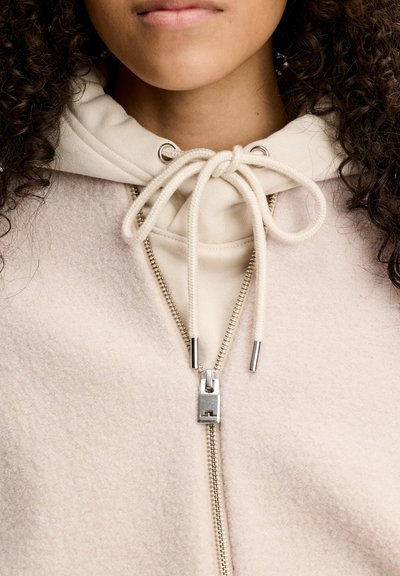 Person wearing a light beige fleece jacket with a silver zipper and an off-white hoodie underneath tied with a bow at the neck.