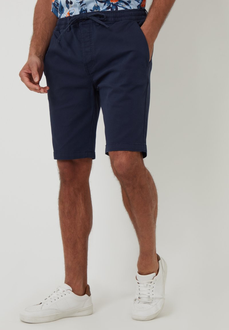 Shorts bleu marine avec une taille élastique et un cordon de serrage, dotés de poches latérales et d'un tissu en coton lisse. Portés avec des baskets blanches.