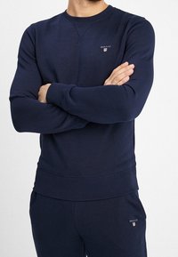 Man som bär en marinblå GANT-tröja och matchande byxor med armarna i kors mot en enfärgad vit bakgrund.