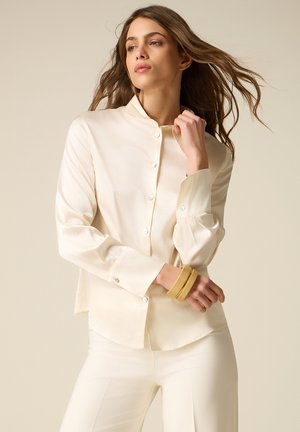 Jonge vrouw met golvend bruin haar, gekleed in een crèmekleurig overhemd met knopen en bijpassende broek, geaccentueerd met twee gele armbanden.