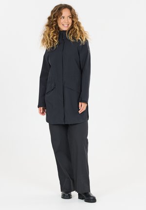 Femme aux cheveux bouclés portant un long manteau noir, un pantalon large noir et des bottes noires, debout et souriante, regardant légèrement sur le côté.