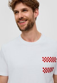 Camiseta de algodón blanca con un bolsillo en el pecho con un patrón de cuadros rojos y blancos. Cuello redondo y mangas cortas. Textura suave.