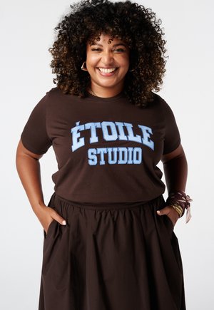 Femme souriante aux cheveux bouclés porte une chemise marron foncé avec "ÉTOILE STUDIO" en bleu clair et une jupe assortie, mains dans les poches.