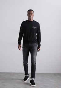 Suéter negro de manga larga con un pequeño logo bordado en el pecho, combinado con jeans gris oscuro y zapatillas negras. Diseño minimalista.