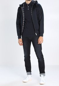 Navy zip-up jacket with plaid lining, black polo shirt, dark jeans, and white sneakers. Focus on smooth fabric and casual fit.