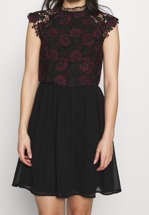 Sukienka bez rękawów z czarną spódnicą i czarną koronkową górą ozdobioną ciemnoczerwonym haftem kwiatowym oraz koronkowymi krawędziami z falbanką na ramionach.
